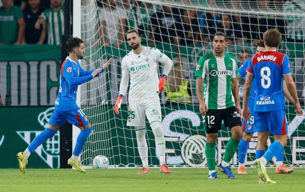 Dynamic soccer match with players competing near the goal, featuring team jerseys, goalposts, and a lively crowd in the background.