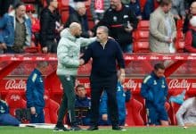 Handshake between two soccer coaches on the sideline of a football match, with team staff and spectators in the background.