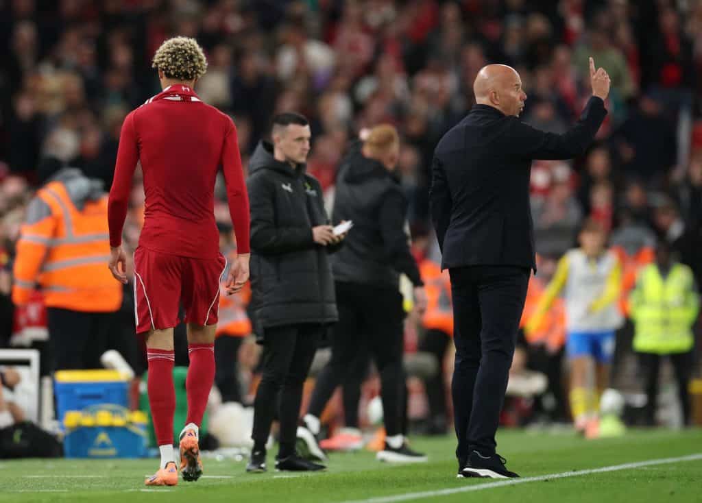 Summer Strikers: Who’s Scoring and Who’s Struggling? Player in red jersey walking on football field during match, coach giving instructions, team staff and crowd in background.