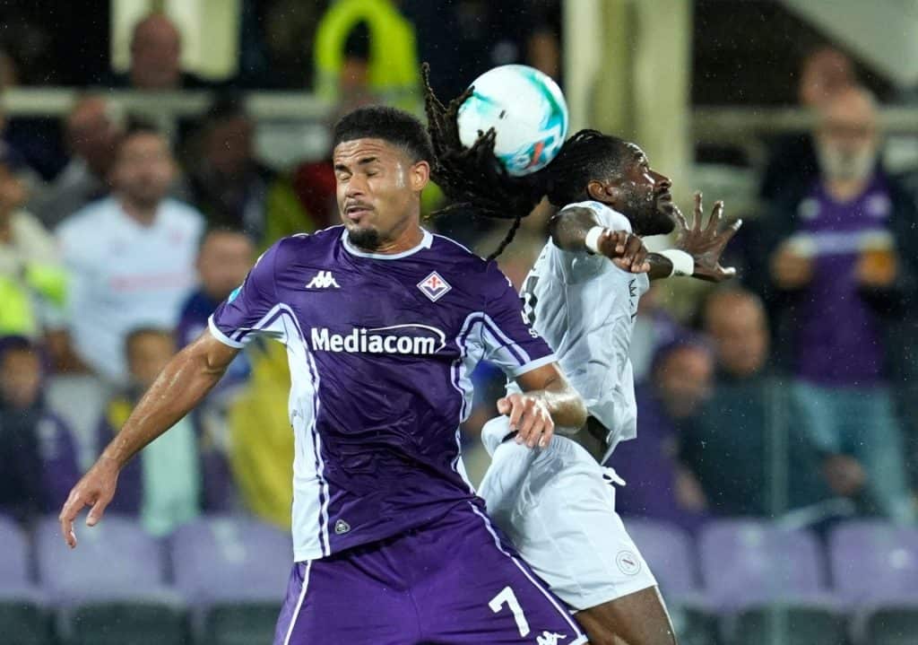 Vibrant soccer action with two players competing for the ball during a high-energy match, showcasing athleticism and intensity. Fans visible in the background under stadium lights.