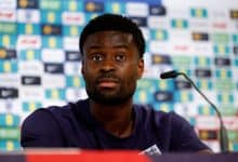 African male athlete at sports press conference, wearing team jersey, with sponsor logos in background.