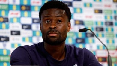 African male athlete at sports press conference, wearing team jersey, with sponsor logos in background.