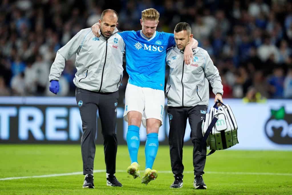 Injured football player being assisted off the field by medical staff during a match, with fans in the background.