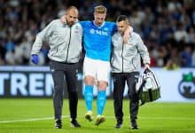 Injured football player being assisted off the field by medical staff during a match, with fans in the background.