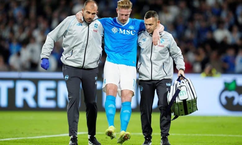 Injured football player being assisted off the field by medical staff during a match, with fans in the background.