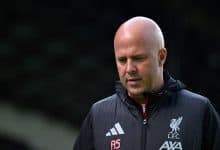 Bald football coach in black sports jacket with Liverpool FC logo, outdoors, focused expression, team management, sports training, leadership, athletic apparel, professional coach, sports apparel, coaching staff, sports performance, soccer training, team strategy, football training, sport coaching, athlete development, soccer team, sports leadership, athletic coaching, sports management, football club, sports professionalism, team sports, coaching expertise, athlete mentoring, soccer tactics, sports motivation, team unity, football tactics, sports discipline, player development, athletic success, sports passion, coaching career, sports excellence, competitive sports, soccer game, sports background, athletic discipline.