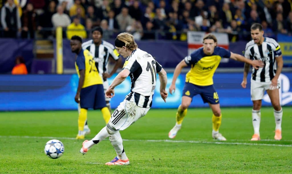 A soccer player in a black and white striped uniform kicking a ball during a match, with opponents in yellow and blue jerseys in the background at a stadium.