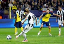 A soccer player in a black and white striped uniform kicking a ball during a match, with opponents in yellow and blue jerseys in the background at a stadium.