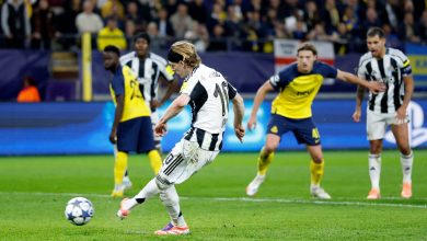 A soccer player in a black and white striped uniform kicking a ball during a match, with opponents in yellow and blue jerseys in the background at a stadium.
