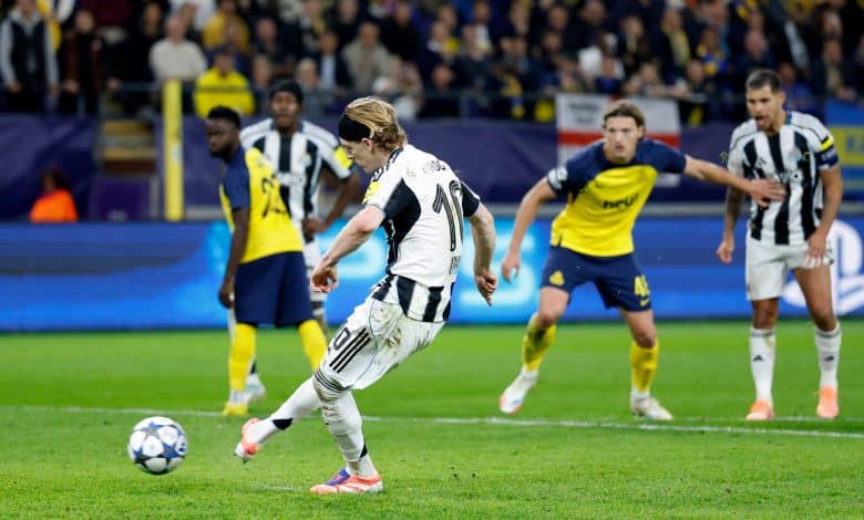 A soccer player in a black and white striped uniform kicking a ball during a match, with opponents in yellow and blue jerseys in the background at a stadium.