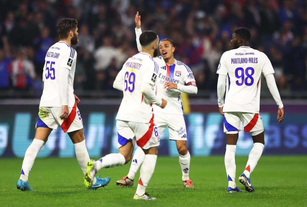 Vibrant soccer match scene featuring players in white and blue kits celebrating on the field, with a blurred crowd in the background, highlighting competitive sports action.