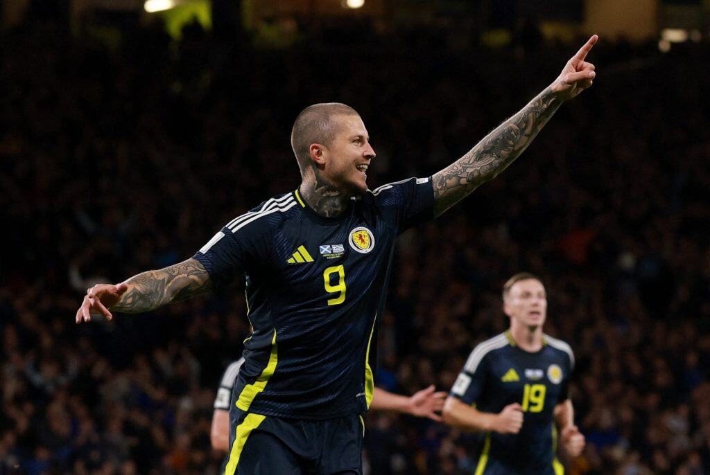 European Night of Goals: Denmark Dominate, Scotland Rise, Faroe Islands Shock Everyone Vibrant image of a football player celebrating a goal during a match, wearing a dark blue jersey with yellow accents and the number 9, with arms raised and tattoos visible, stadium filled with fans in the background.