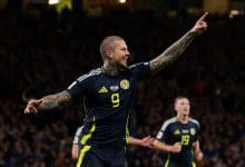 Vibrant image of a football player celebrating a goal during a match, wearing a dark blue jersey with yellow accents and the number 9, with arms raised and tattoos visible, stadium filled with fans in the background.