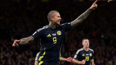 Vibrant image of a football player celebrating a goal during a match, wearing a dark blue jersey with yellow accents and the number 9, with arms raised and tattoos visible, stadium filled with fans in the background.
