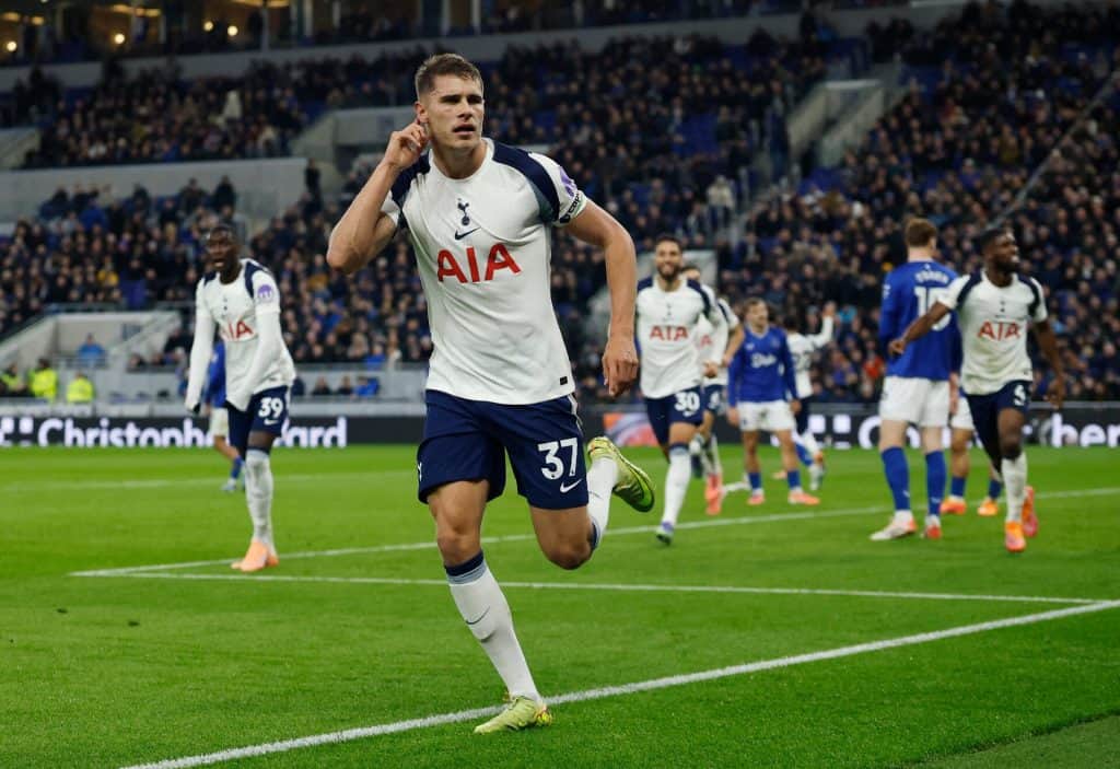 Energetic football player celebrating during a match with team members on the field, wearing Tottenham Hotspur kit, under stadium lights, with a cheering crowd in the background.