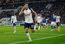 Energetic football player celebrating during a match with team members on the field, wearing Tottenham Hotspur kit, under stadium lights, with a cheering crowd in the background.