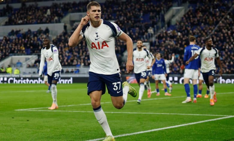Energetic football player celebrating during a match with team members on the field, wearing Tottenham Hotspur kit, under stadium lights, with a cheering crowd in the background.