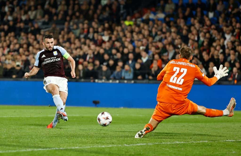 Underdogs Steal the Show on a Wild Europa League Night Aston Villa player in a black and blue kit and goalkeeper in orange uniform during a football match with a crowded stadium in the background.