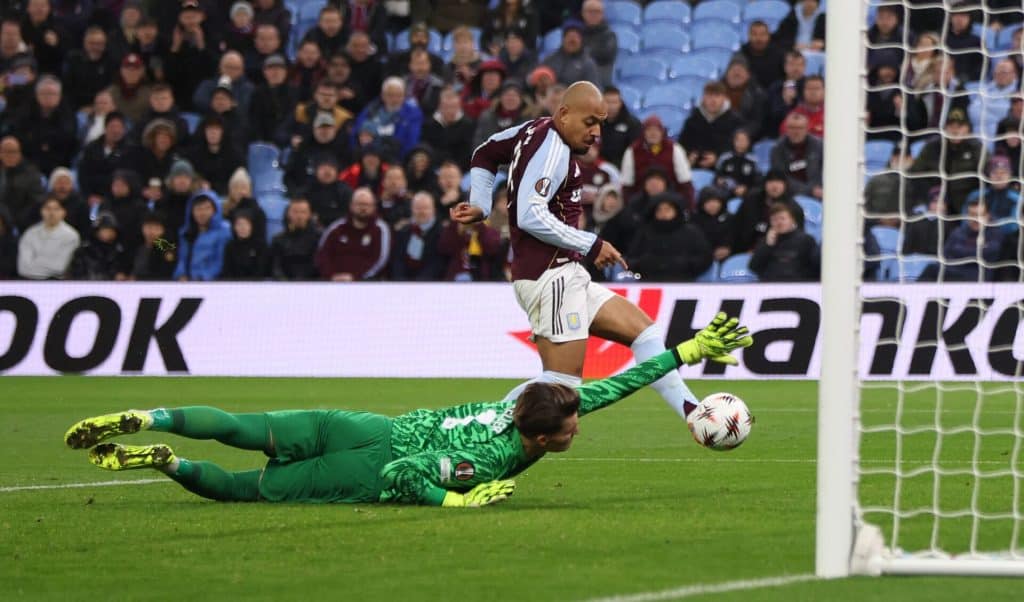 Goalkeeper in green diving to save ball during match, player in maroon jersey shooting towards goal, stadium filled with spectators, sports action, football match scene.