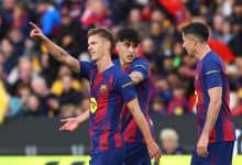 Young soccer players in FC Barcelona uniforms celebrating a goal during a match, with a lively stadium crowd in the background.