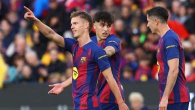 Young soccer players in FC Barcelona uniforms celebrating a goal during a match, with a lively stadium crowd in the background.