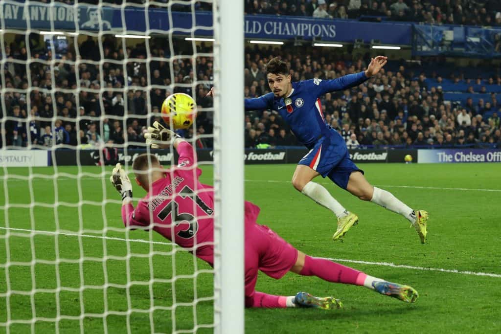 1. Soccer player in Chelsea kit scoring a goal past goalkeeper in pink goalkeeper uniform during match at Stamford Bridge stadium.