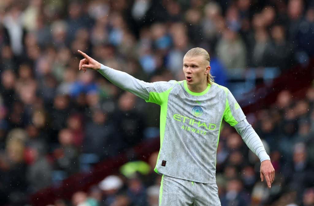 Young male football player wearing Manchester City kit, pointing and shouting on the field during a match, with a crowd in the background.