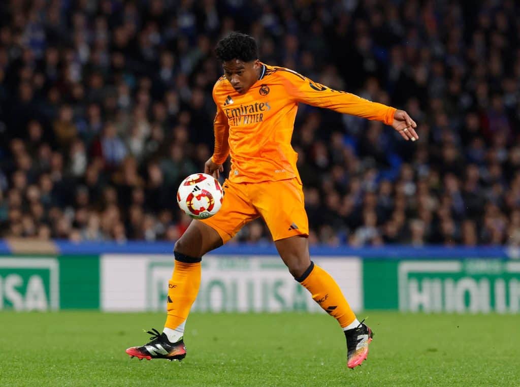 Athlete in bright orange soccer uniform with "Emirates" sponsor, executing a skillful move during a professional match on a green field.