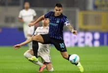 A soccer player in black and blue jersey controlling the ball during a match, with an opponent attempting to intercept, on a green grass field under stadium lights.