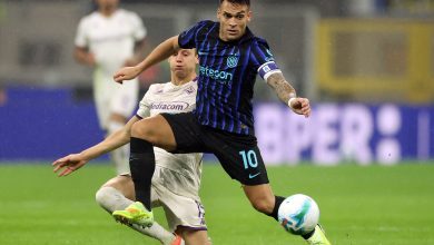 A soccer player in black and blue jersey controlling the ball during a match, with an opponent attempting to intercept, on a green grass field under stadium lights.