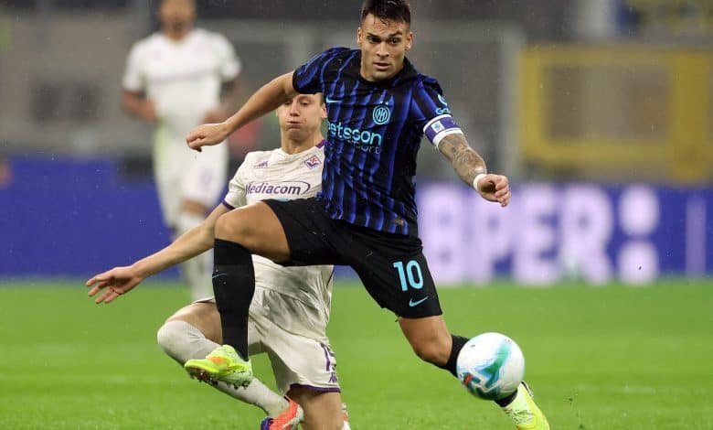 A soccer player in black and blue jersey controlling the ball during a match, with an opponent attempting to intercept, on a green grass field under stadium lights.