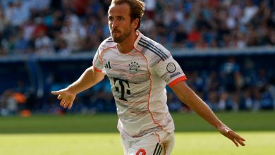 Dynamic soccer player celebrating on the field during match, wearing Bayern Munich kit, in outdoor stadium, with a focused expression and extended arm, under bright sunlight.