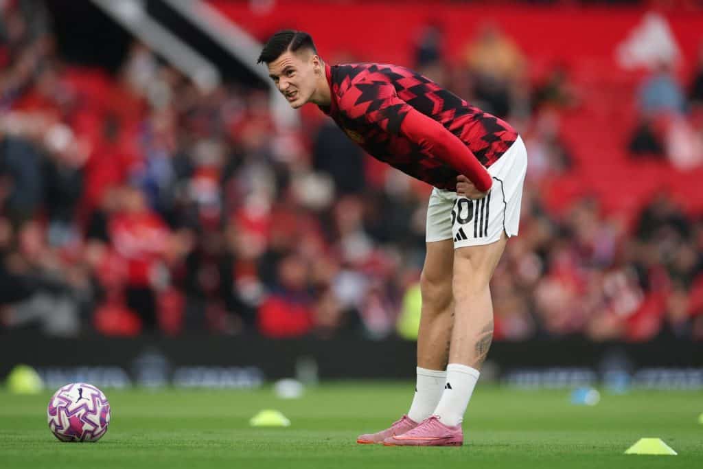 Young male soccer player bending over on field during training or warm-up, wearing red and black jersey, white shorts, pink soccer cleats, preparing for match, stadium atmosphere.