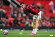 Young male soccer player bending over on field during training or warm-up, wearing red and black jersey, white shorts, pink soccer cleats, preparing for match, stadium atmosphere.