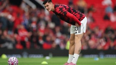 Young male soccer player bending over on field during training or warm-up, wearing red and black jersey, white shorts, pink soccer cleats, preparing for match, stadium atmosphere.