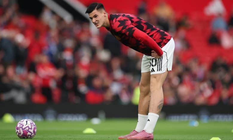 Young male soccer player bending over on field during training or warm-up, wearing red and black jersey, white shorts, pink soccer cleats, preparing for match, stadium atmosphere.