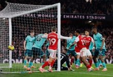 A football match in progress at Emirates Stadium with players from two teams competing for goal opportunity, with spectators in the background.