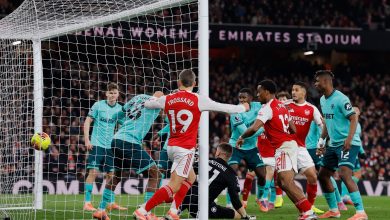 A football match in progress at Emirates Stadium with players from two teams competing for goal opportunity, with spectators in the background.