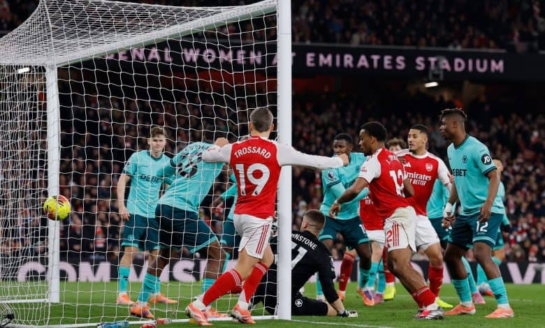A football match in progress at Emirates Stadium with players from two teams competing for goal opportunity, with spectators in the background.