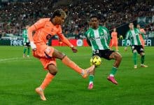 Soccer players competing on the field during a competitive match; one in orange goalkeeper gear and the other in green, focused on the ball.