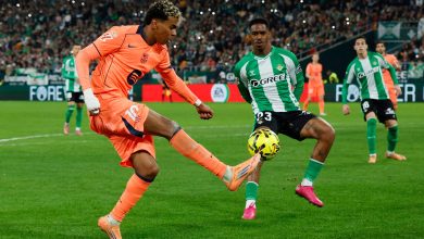 Soccer players competing on the field during a competitive match; one in orange goalkeeper gear and the other in green, focused on the ball.