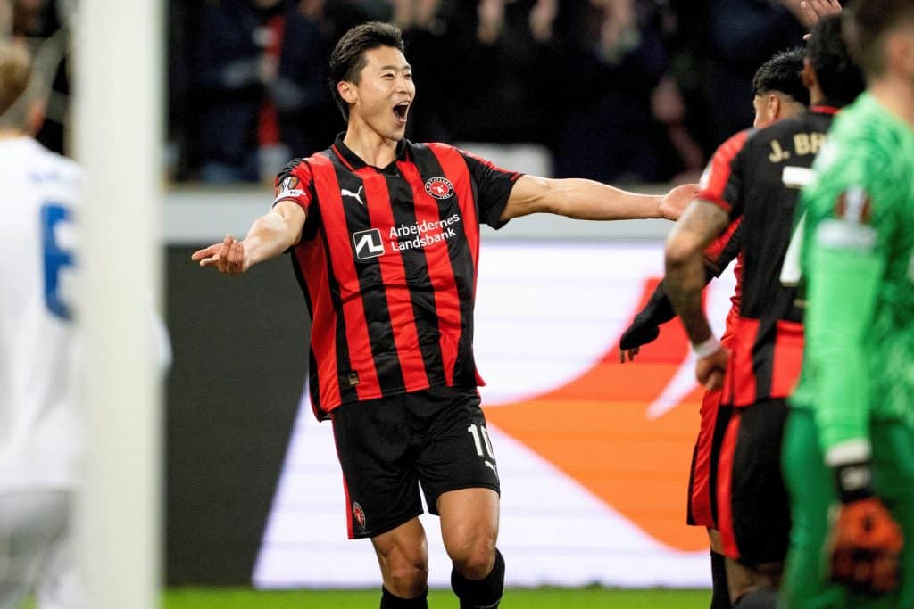 A soccer player in a black and red kit passionately celebrates a goal, surrounded by teammates and opponents in a stadium, highlighting dynamic football action at ZareSport.et.
