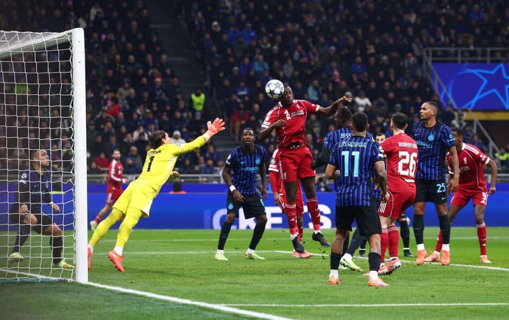 Soccer players jump for a header during a competitive match, with the goalkeeper reaching out and fans watching intently in the stadium, showcasing the excitement of professional football.
