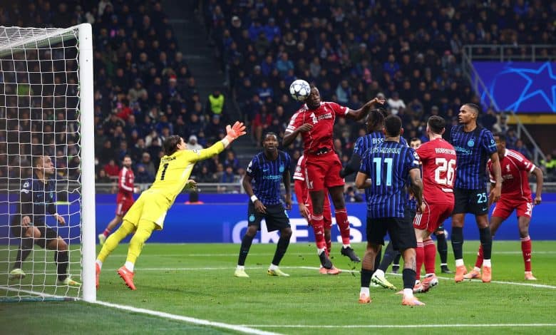Soccer players jump for a header during a competitive match, with the goalkeeper reaching out and fans watching intently in the stadium, showcasing the excitement of professional football.