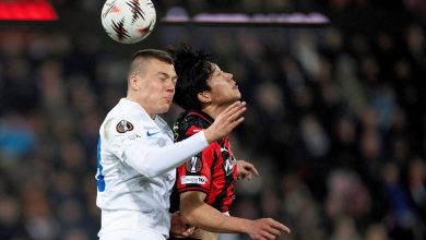 A soccer match between two players vying for the ball during an intense game. One player in a white jersey and the other in black and red, both concentrating on the ball mid-air under stadium lights.