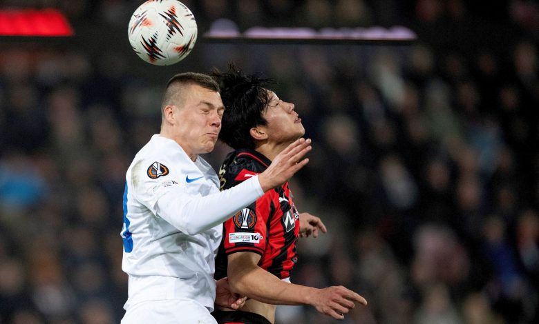 A soccer match between two players vying for the ball during an intense game. One player in a white jersey and the other in black and red, both concentrating on the ball mid-air under stadium lights.