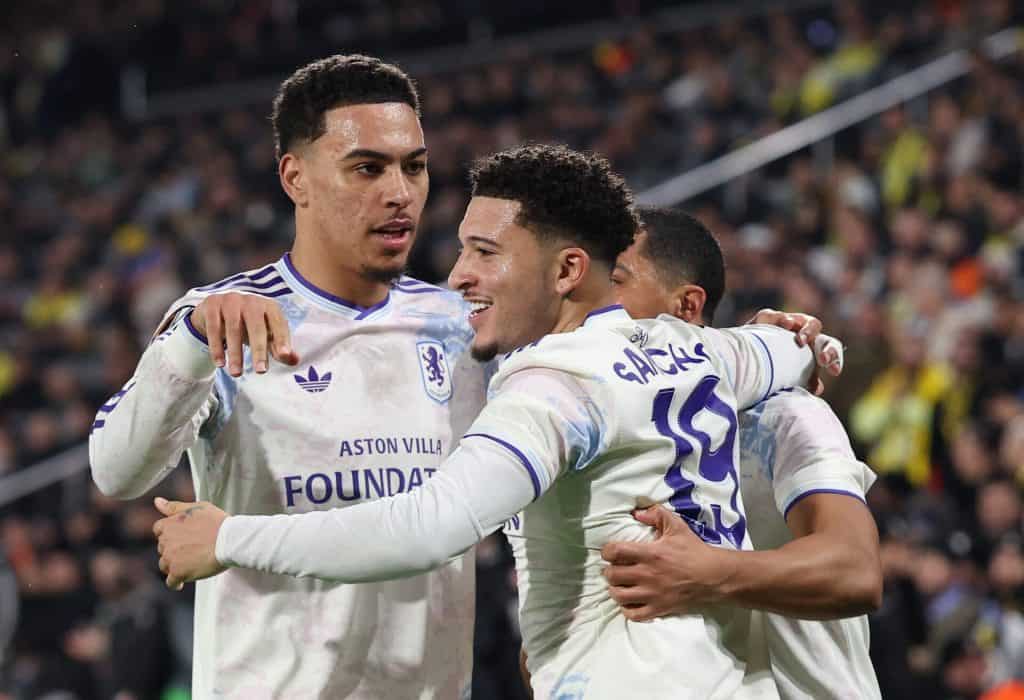 Soccer players from Aston Villa celebrate together on the field during a match, displaying excitement and teamwork. The players are wearing white kits with purple accents, amid a vibrant crowd in the background.