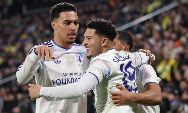 Soccer players from Aston Villa celebrate together on the field during a match, displaying excitement and teamwork. The players are wearing white kits with purple accents, amid a vibrant crowd in the background.
