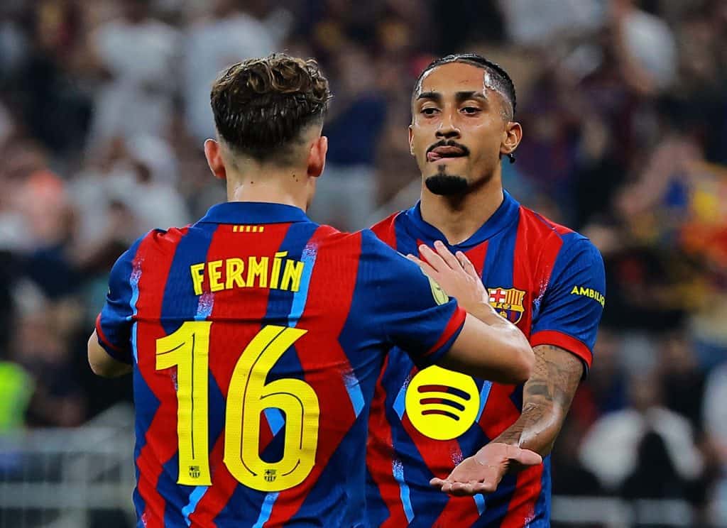 A close-up of two soccer players in Barcelona jerseys, one with "Fermin" and number 16, celebrating a goal on the field. The intense match atmosphere and team camaraderie are captured, emphasizing soccer excitement and sportsmanship.