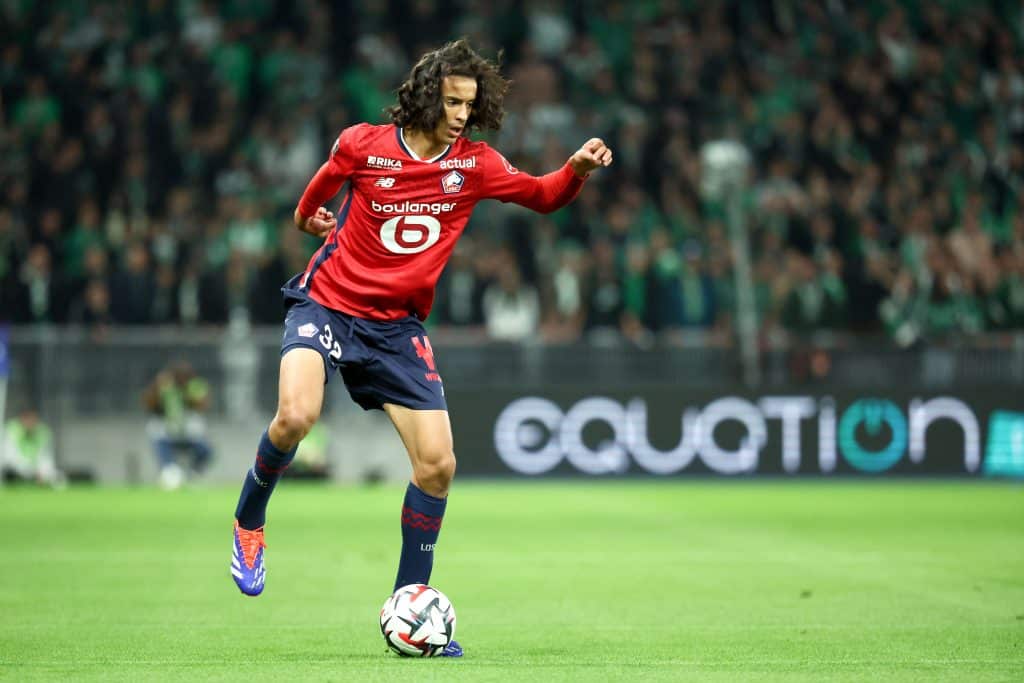 Athletic soccer player wearing red jersey and navy shorts dribbling ball during match with crowd in background.